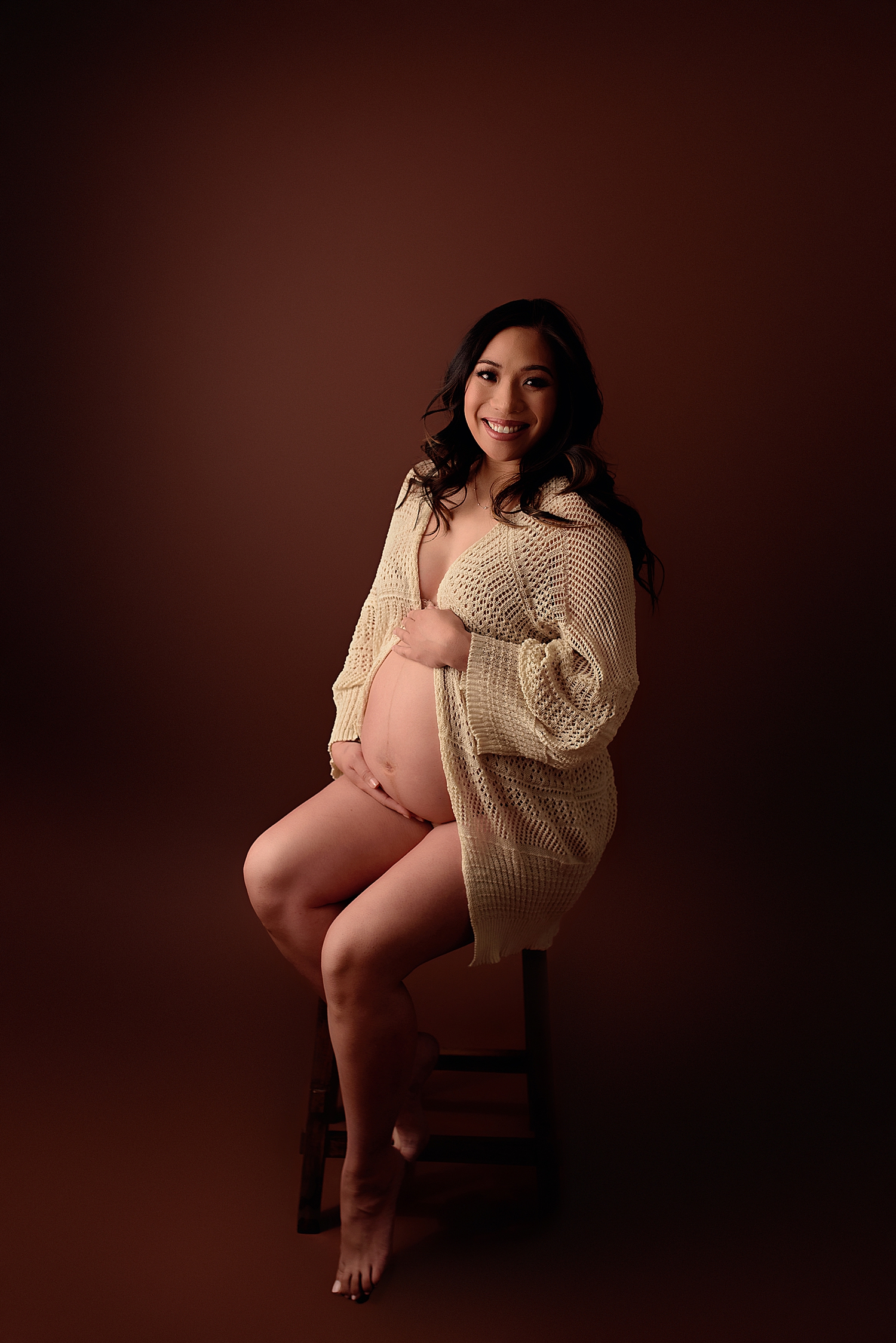 pregnant person in cream knit sweater on studio stool by Yaffa Koff Photography